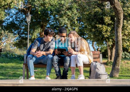 Eine Gruppe von Freunden im Teenageralter, die auf einem Tablet im Park auf einer Bank sitzen Stockfoto