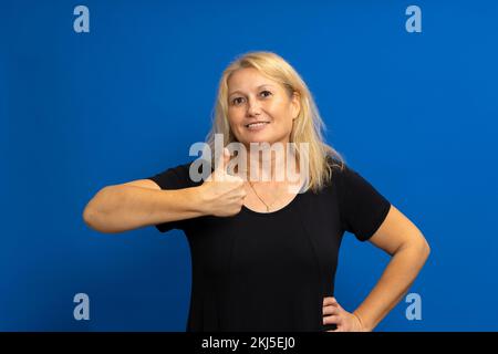 Schöne weiße blonde Frau, die ein schwarzes T-Shirt über einem isolierten blauen Hintergrund trägt und mit der Hand eine fröhliche Daumen-hoch-Geste macht. Genehmigungsausdruck Stockfoto