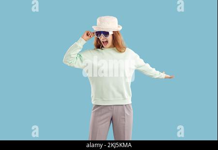 Lustige, fröhliche junge Frau mit Hut, Sonnenbrille, Minze-Baumwolle-Sweatshirt, das auf blauem Hintergrund tanzt. Stockfoto