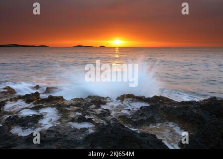 Bild des Sonnenaufgangs mit Wellen auf den Felsen am Strand von Bombay, Santa Eulalia, Ibiza, Spanien. Stockfoto