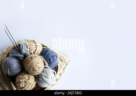 Graue, braune und blaue Strickfäden in einem beigen Korb mit Stricknadeln und einem Stapel Strickmützen für Winter und Herbst, Strickkochfeld Stockfoto
