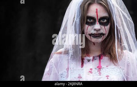 Porträt einer Zombie-Frau im Hochzeitskleid mit Schleier und Bühnenschminke, die in die Kamera schaut. Das Mädchen hat weißes Make-up und Blutflecken auf ihr Gesicht aufgetragen Stockfoto