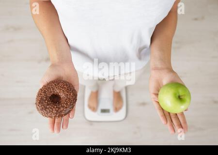 Eine Frau mit einer Badezimmerwaage, die einen Apfel und einen Donut in der Hand hält Stockfoto