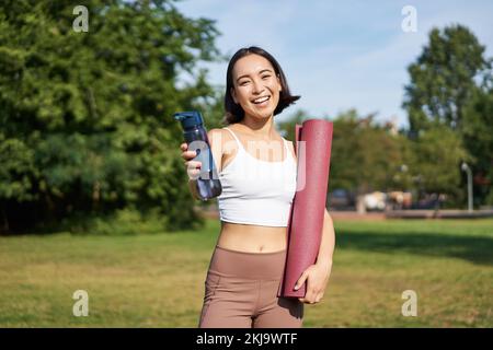 Porträt eines glücklichen asiatischen Mädchens, eine Fitnessfrau, die dir nach dem Training eine Flasche Wasser zum Trinken gibt, mit einer Gummimatte für Übungen im Park Stockfoto