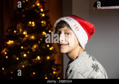 Ein Junge lächelt neben dem Weihnachtsbaum mit einem Weihnachtsmütze, grünen Augen, Lächeln. Der Junge feiert weihnachten in Kolumbien neben dem weihnachtsbaum Stockfoto