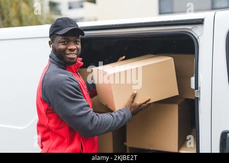 Schwarzer junger Erwachsenenlieferant in Arbeitskleidung und schwarze Mütze, die Pakete aus dem weißen Lieferwagen auslädt und in die Kamera lächelt. Horizontale Außenaufnahme. Hochwertiges Foto Stockfoto