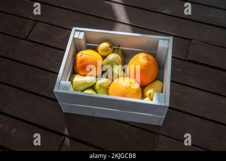 Eine Holzkiste gefüllt mit reifen Zitrusfrüchten und verschiedenen Birnen, die in den Sonnenstrahlen der Morgensonne auf einer Oberfläche von Akazienplanken gebadet wurden Stockfoto