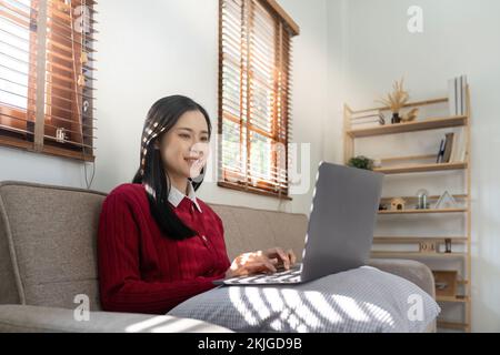 Junge asiatische Frau, die einen Laptop benutzt und im Wohnzimmer auf den Bildschirm schaut, Remote Job oder Arbeit von zu Hause Konzept. Stockfoto