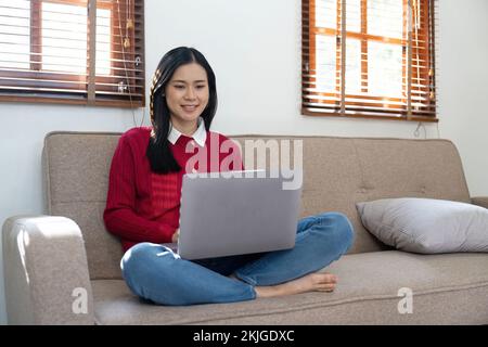 Junge asiatische Frau, die einen Laptop benutzt und im Wohnzimmer auf den Bildschirm schaut, Remote Job oder Arbeit von zu Hause Konzept. Stockfoto