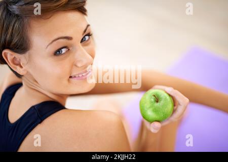 Ihre Geheimwaffe für ein gesundes Leben. Eine sportliche junge Frau, die auf dem Sportfußboden sitzt und einen Apfel isst. Stockfoto