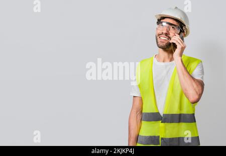 Lächelnder Ingenieur, der am Telefon telefoniert, isoliert mit Kopierraum. Junger Bauingenieur in Weste, der auf dem Handy spricht, isoliert. Bauingenieurkonzept Stockfoto