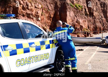 Ein Mann der Küstenwache, der durch ein Fernglas an den Klippen in Pennington Point, Sidmouth, Devon, Großbritannien, schaut. Stockfoto