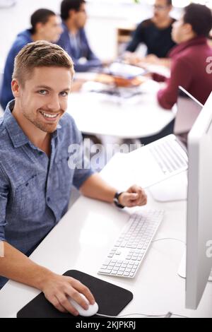 Wie unsere Software sind auch unsere Programmierer benutzerfreundlich. Porträt eines glücklichen jungen Mannes, der an seinem Computer in einem Büro arbeitet. Stockfoto