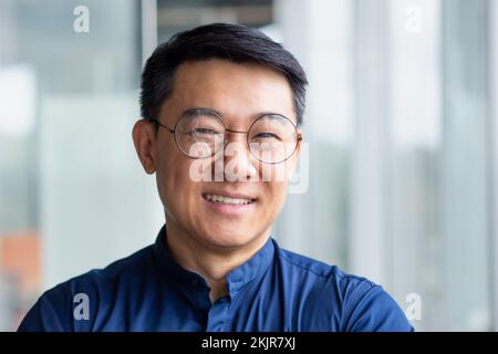 Nahaufnahme Foto eines erfolgreichen und glücklichen asiatischen Geschäftsmanns, eines reifen Chefs mit Brille, der im modernen Bürogebäude arbeitet und lächelt und in die Kamera schaut Stockfoto