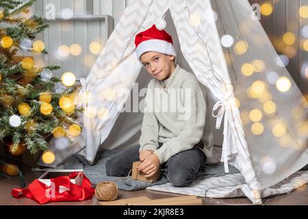 Familienweihnachtsfest. Kinder öffnen Weihnachtsgeschenke. Ein Teenager mit Weihnachtsmannmütze öffnet ein Geschenk und lächelt, während er auf dem Boden des Hauses sitzt Stockfoto