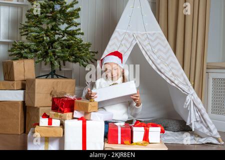 Ein emotionales Mädchen in einem Weihnachtsmütze sitzt auf dem Boden neben einem Stapel ihrer Geschenke im Kinderzimmer. Das Kind ist glücklich mit Geschenken vom Weihnachtsmann. Das Chi Stockfoto