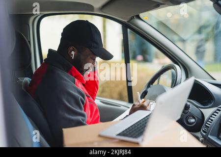 Schwarzer junger Erwachsener, der auf dem Fahrersitz des Lieferwagens sitzt, Dokumente ausfüllt und an einem Laptop arbeitet. Horizontale Aufnahme. Hochwertiges Foto Stockfoto