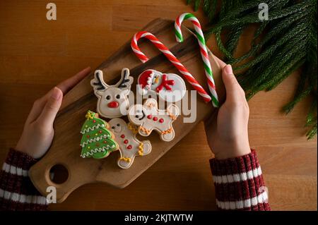 Die Hände einer Frau halten ein hölzernes Tablett mit süßem und leckerem Lebkuchen und Süßigkeiten auf einem Holztisch. Blick von oben, traditionelle Weihnachtsbonbons Stockfoto