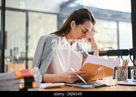 Gestresste asiatische Geschäftsfrau, die sich nach dem Lesen eines Dokuments verärgert und deprimiert fühlt, während sie an ihrem Schreibtisch arbeitet und ein Kündigungsschreiben erhält Stockfoto