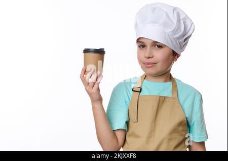 Ein hübscher Teenager mit Kochmütze und beigefarbener Schürze, der einen Kaffee zum Mitnehmen in umweltfreundlichen Pappbechern verkauft Stockfoto