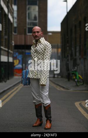 Ein cooler, stylischer Mann in einer Londoner Straße, der Cowboystiefel, ein gepunktetes Hemd und eine Männertasche trägt Stockfoto