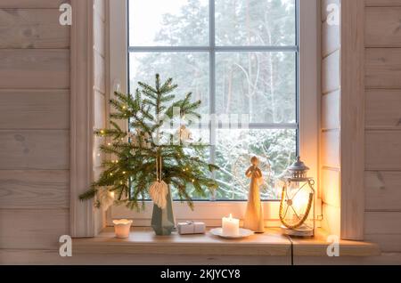 Weihnachtslaterne, Weihnachtsengel, Weihnachtsbaum und weiße Einrichtung am Fenster eines Holzhauses Stockfoto