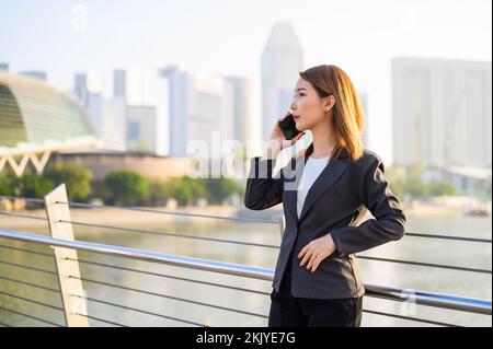Junge asiatische Geschäftsfrau mit Smartphone, die im Finanzviertel der modernen Stadt posiert. Geschäfts- und Kommunikationskonzept Stockfoto
