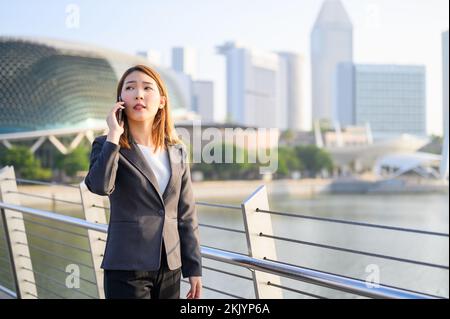 Junge asiatische Geschäftsfrau mit Telefon im Freien mit Smartphone im modernen Finanzviertel der Stadt stehen. Business- und Kommunikationskonzept Stockfoto