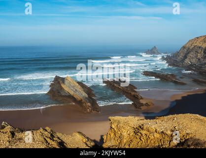 Blick auf den leeren Sandstrand von Alteirinhos mit Meereswellen, Klippen, Steinen und Felsen im morgendlichen goldenen Licht an der wilden Küste von Rota Vicentina, Odemira Stockfoto