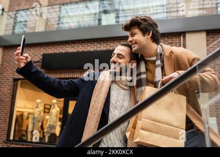 Fröhliches schwules Paar in trendigen Mänteln und Schals Selfie auf dem Smartphone auf der Rolltreppe, Stockbild Stockfoto