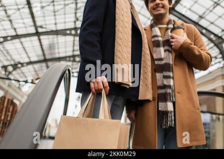 Kurze Aussicht auf einen trendigen schwulen Mann mit Einkaufstüten in der Nähe eines verschwommenen Freundes mit einem Getränk zum Mitnehmen auf der Rolltreppe, Bild der Aktien Stockfoto