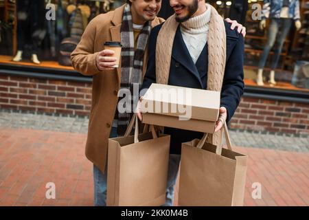 Kurze Aussicht auf einen lächelnden schwulen Mann mit Kaffee zum Mitnehmen, einen Freund umarmen mit Schuhkarton und Einkaufstaschen auf der Straße, Bild der Börse Stockfoto