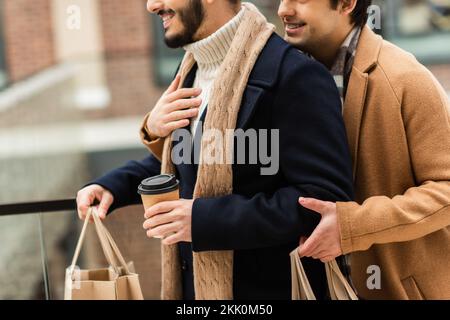 Kurze Aussicht auf einen lächelnden schwulen Mann, der einen bärtigen Freund umarmt, mit Kaffee zum Mitnehmen und Einkaufstaschen, Stockbild Stockfoto