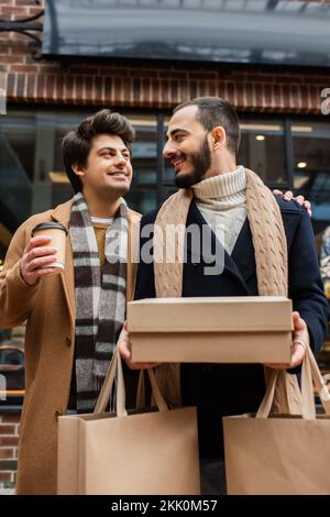 Glückliches schwules Paar in trendigen Outfits, das sich anschaut, während es mit Käufen und Getränken zum Mitnehmen steht, Stock Image Stockfoto