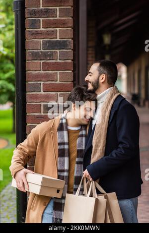 Glücklicher schwuler Mann mit Zahnspangen und geschlossenen Augen, der einen Schuhkarton hielt, während er sich auf einen bärtigen Freund mit Einkaufstaschen stützte, Bild der Aktie Stockfoto