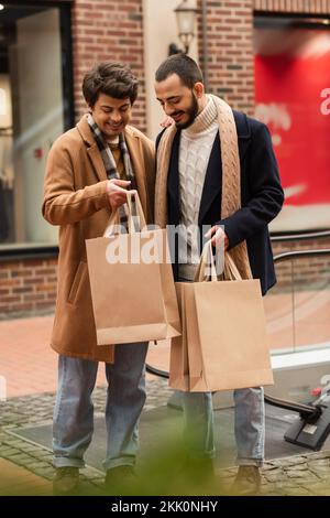 Fröhliches schwules Paar in trendigen Kleidern mit Einkaufstaschen in der Nähe eines verschwommenen Gebäudes, Bild der Aktien Stockfoto