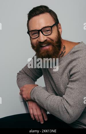 Porträt eines positiven Mannes mit Brille und Jumper, der auf einem Stuhl sitzt, isoliert auf einem grauen Stockbild Stockfoto