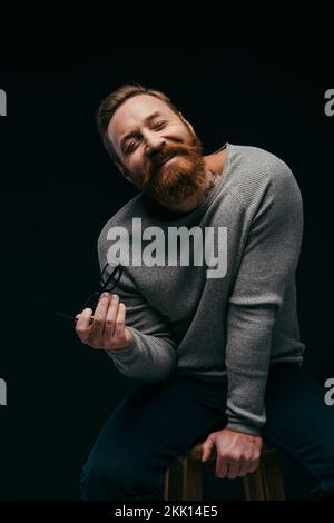 Lächelnder bärtiger Mann im Jumper, der eine Brille hält, während er auf dem Stuhl sitzt, isoliert auf schwarzem Stockbild Stockfoto