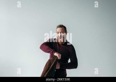 Ein verblüffter und bärtiger Mann im Rollkragenpullover hält einen Blazer und schaut auf die Kamera isoliert auf einem grauen Stockbild Stockfoto