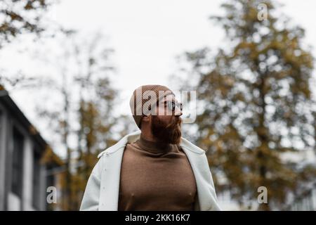 Stilvoller Mann mit Bart, Mütze und trendiger Sonnenbrille mit Blick auf Bäume und bewölkten Himmel, Stockbild Stockfoto