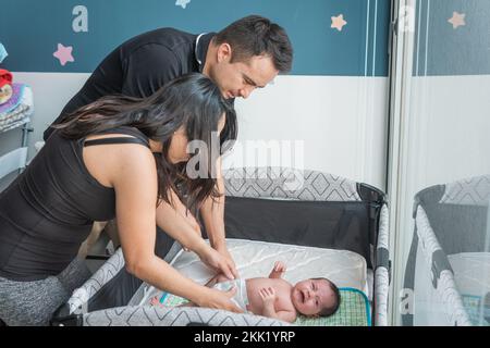 Vater und Mutter aus Latino wechseln die Windel, bis sie ihr weinendes Baby im Kinderbett haben Stockfoto