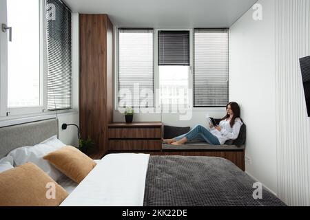 Frau liest Buch in einem Schlafzimmer mit Bett und Holzgarderobe in einem modernen Apartment Stockfoto