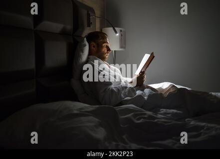Weißer Mann im mittleren Alter, der sich im Bett entspannt und ein Bestseller-Buch liest, mit eingeschalteter Nachttischlampe. Abendliche Entspannung, Hobbys, freie Zeit conce Stockfoto