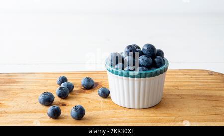 Nahaufnahme einer kleinen weißen Keramikschüssel gefüllt mit frischen Blaubeeren. Auf einem Holztisch platziert Stockfoto