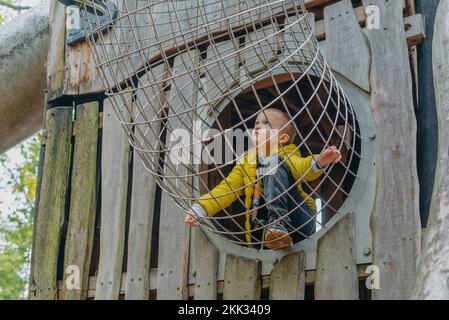 Ein Kind klettert an einem heißen Sommertag in einem Park auf einem Spielplatz auf ein Berggitter. Kinderspielplatz in einem öffentlichen Park, Unterhaltung und Erholung Stockfoto