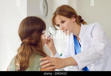 Professionelle Dermatologin untersucht das Gesicht eines Teenagers mit einer Lupe in ihrem Büro. Stockfoto