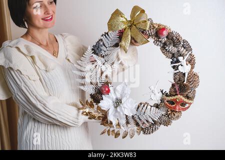 Nahaufnahme Frau hängt wunderschönen Weihnachtskranz. Die Basis des Kranzes sind Fichtenäste, keine verschwenderische Dekoration in weiblichen Händen Stockfoto