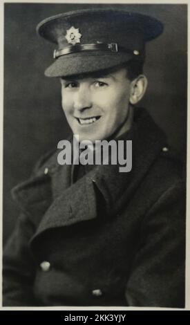 Britischer Soldat in Uniform, 2. Weltkrieg. Gekleidet in einem tollen Mantel und einer schicken Mütze mit Militärabzeichen, die in die Kamera lächelt. Hübscher junger Junge. In einem Porträtstudio aufgenommen und für die Familie auf eine Postkarte gedruckt. Schwarzweißfoto. Unbekannte Person. Stockfoto