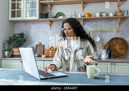 Eine junge hispanische Freiberuflerin in Brillenarbeit kann zu Hause nicht mit einem Laptop arbeiten. Die Lichter sind aus, die Termine sind an. Sie sitzt verwirrt und wütend in der Küche am Tisch und spreizt ihre Hände. Stockfoto