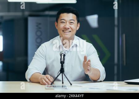 Porträt eines jungen Asiaten, der seinen Audio-Podcast mit einem Mikrofon in seinem kleinen Fernsehstudio ausstrahlt. Er sieht in die Kamera und lächelt. Stockfoto